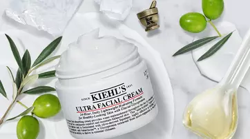 A jar of Kiehl's Ultra Facial Cream surrounded by olive branches, a small bowl of cream, and a decorative spoon on a marble surface.