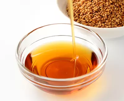 A glass bowl filled with golden-brown sesame oil, with a stream of oil pouring into it from above. In the background, a white bowl contains dry sesame seeds.