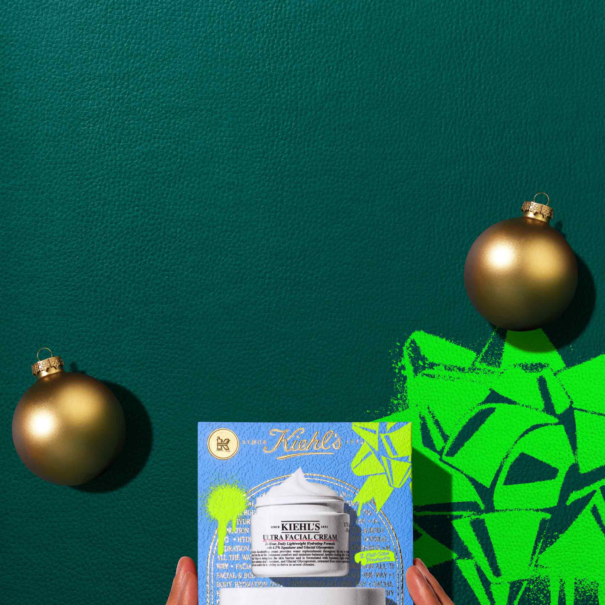 A graphic, set against a dark green background, features two open white jars of Kiehl’s cream at the top, showing visible swirls of product. The smaller jar is "KIEHL’S ULTRA FACIAL CREAM," and the larger jar is "KIEHL’S ULTRA BODY MEGA MOISTURE SQUALANE CREAM." Below them, a blue Kiehl’s gift box displays the same two products. Two gold spherical ornaments are placed on either side of the gift box. The gift box also has bright green graphic paint splatters and a bow design.