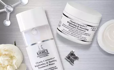 A flat lay image featuring skincare products from Kiehl's: a bottle of Clearly Corrective Brightening & Smoothing Treatment Water and a jar of Clearly Corrective Brightening & Smoothing Moisture Treatment, surrounded by a delicate flower and a silver pin.