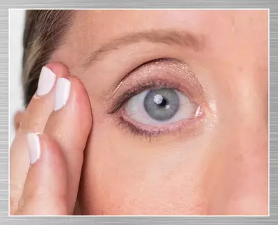 A close-up of a woman's eye, showing her eyelid and skin texture, with her fingers gently touching the outer corner of her eye. She has a subtle makeup look.