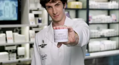 A person in a white lab coat is holding up a jar of Kiehl's Ultra Facial Cream in front of a display of skincare products.