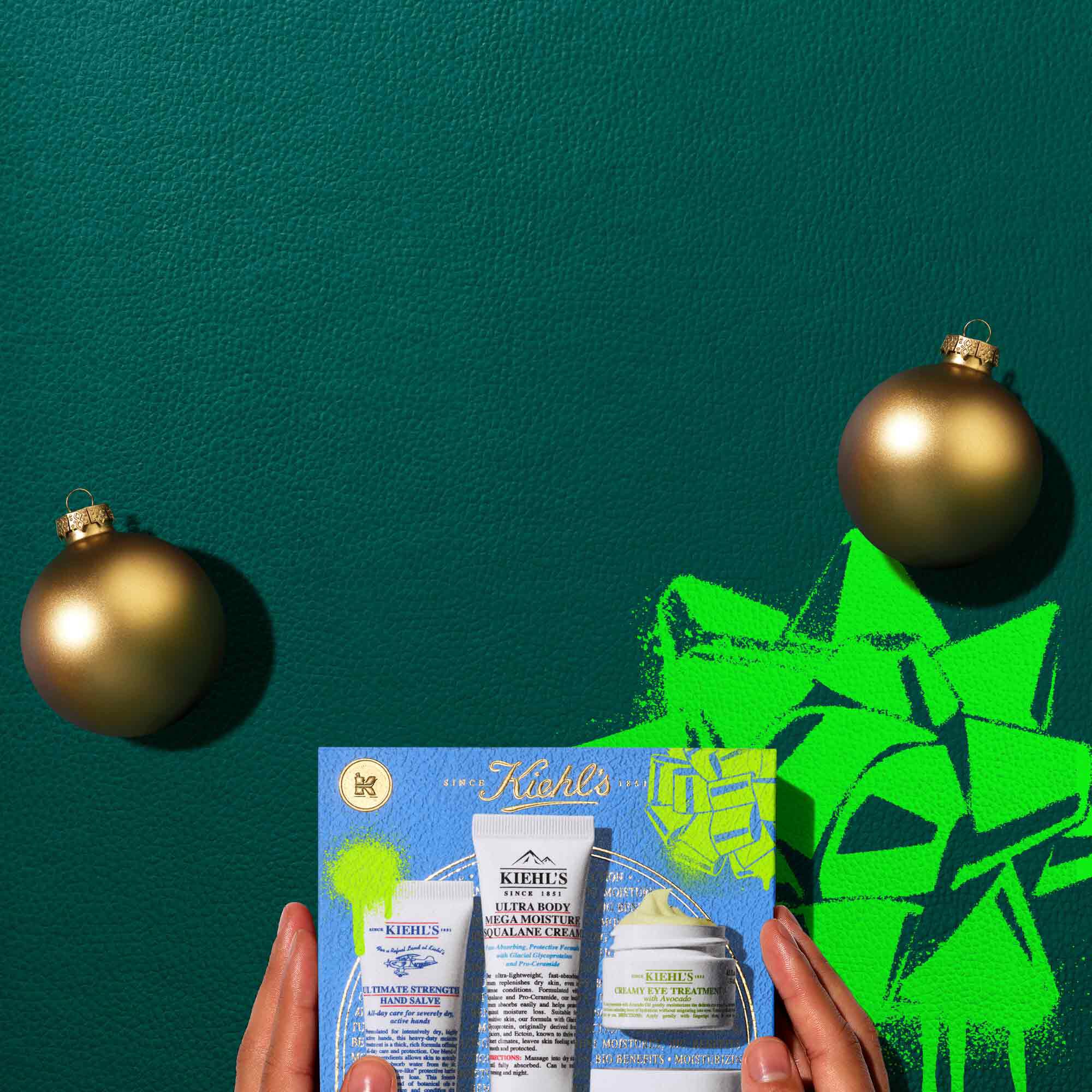 A graphic, set against a dark green textured background, features four Kiehl’s products and a gift box. The top row displays a white tube of "KIEHL’S ULTIMATE STRENGTH HAND SALVE," a white tube of "KIEHL’S ULTRA BODY MEGA MOISTURE SQUALANE CREAM," a small open white jar of "KIEHL’S CREAMY EYE TREATMENT WITH AVOCADO" with visible pale yellow cream, and a white open jar of "KIEHL’S ULTRA FACIAL CREAM" with visible white cream. Two gold spherical ornaments are also present. Below these, a blue Kiehl’s gift box showcases the same four products, decorated with bright green graphic paint splatters and a bow design.