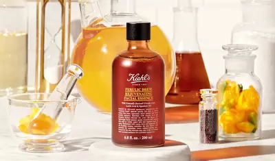 A bottle of Kiehl's Ferulic Brew Rejuvenating Facial Essence is placed on a marble surface, surrounded by various lab glassware filled with colorful liquids and ingredients, including yellow flowers and seeds.