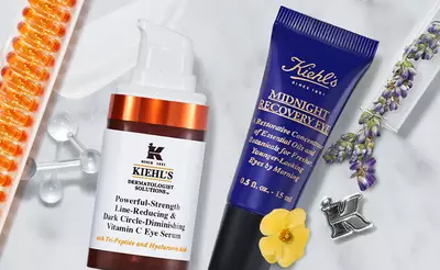 A flat lay of Kiehl's skincare products, featuring a pump bottle of Powerful-Strength Line-Reducing & Dark Circle-Diminishing Vitamin C Eye Serum and a tube of Midnight Recovery Eye cream, surrounded by orange capsules, lavender, and a flower.