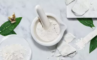 A white mortar and pestle surrounded by loose white powder, broken pieces of powdery substance, and green leaves on a light marble surface.