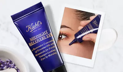 A close-up of a hand applying Kiehl's Midnight Recovery Eye cream under the eye, with a tube of the product and a small dollop of cream on a marble surface in the background.