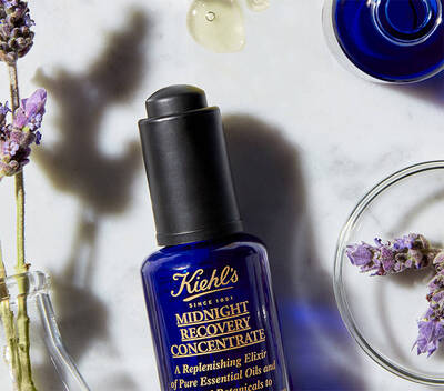 Flacon de ser Kiehl's Midnight Recovery, alături de flori de lavandă și capsule cu uleiuri esențiale pe un fundal marmorat.