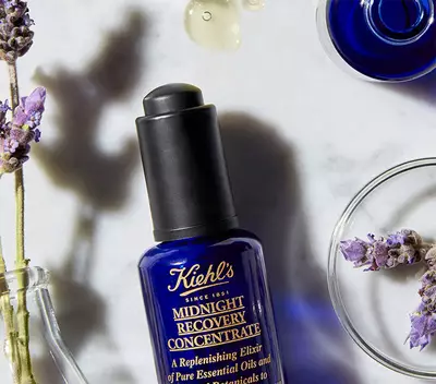 Flacon de ser Kiehl's Midnight Recovery, alături de flori de lavandă și capsule cu uleiuri esențiale pe un fundal marmorat.