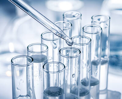 A dropper dispensing liquid into several test tubes filled with clear liquid, set against a blurred laboratory background.