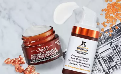A flat lay of Kiehl's skincare products, featuring a jar of Powerful Wrinkle Reducing Eye Cream and a bottle of Powerful-Strength Dark Circle-Diminishing Vitamin C Eye Serum, surrounded by textured elements and orange crystals.